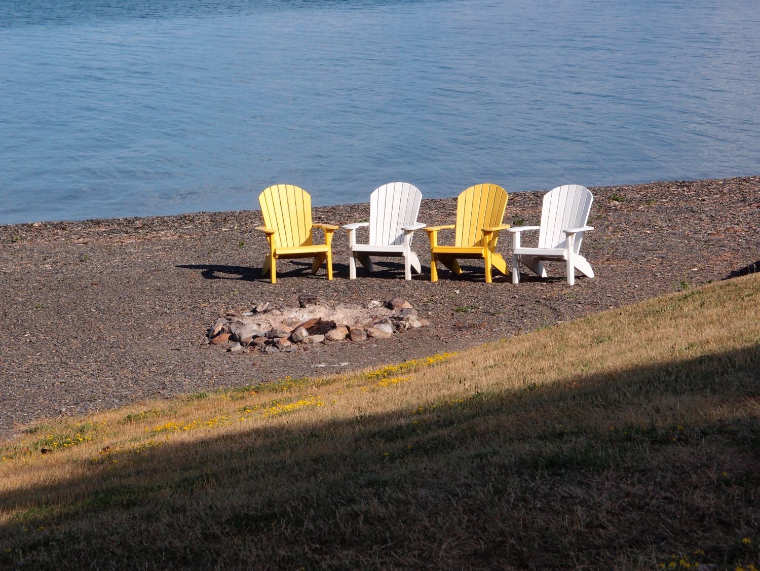 Four Adirondack chairs, alternating yellow and white, facing a fire pit along the shore of Cayuga Lake