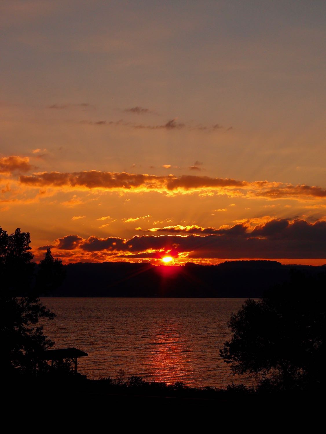 Sunrise over Cayuga Lake, sun between the hills and some clouds with subtle rays radiating outward.