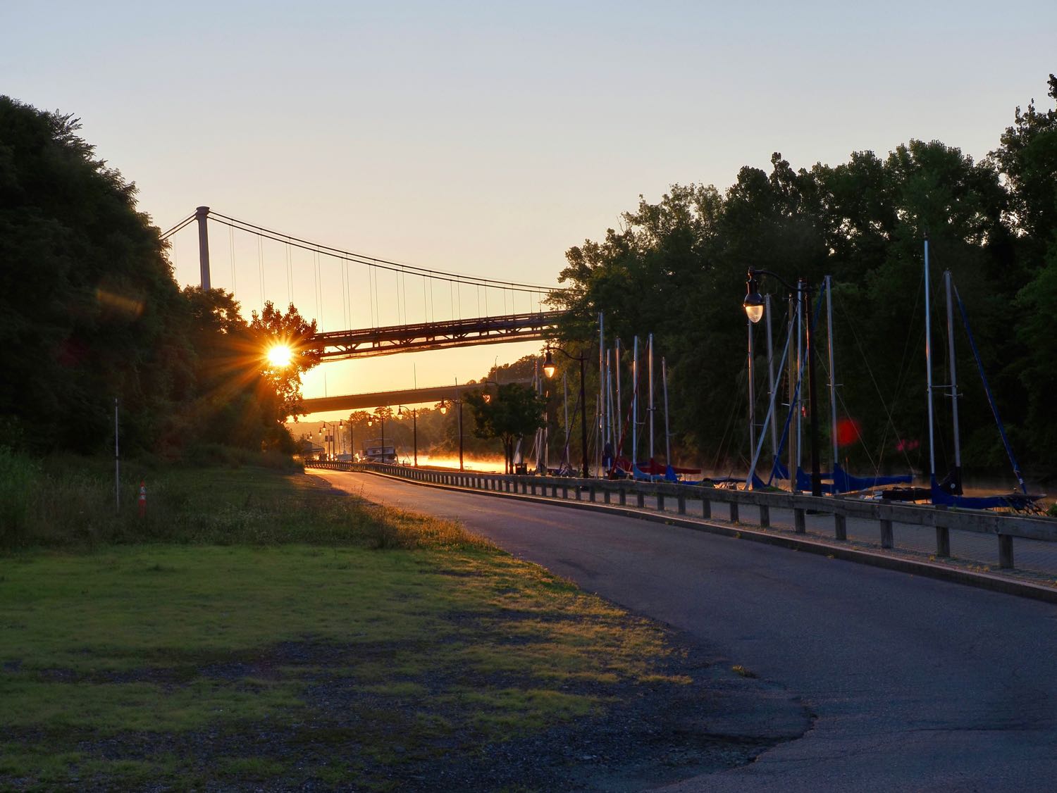 At the Rondout Creek in Kingston NY, looking east toward the sun. Two bridges in the background with a sunstar in the trees. Mist rising from the creek, and a line of sailboat masts and light poles lead toward the bridges.