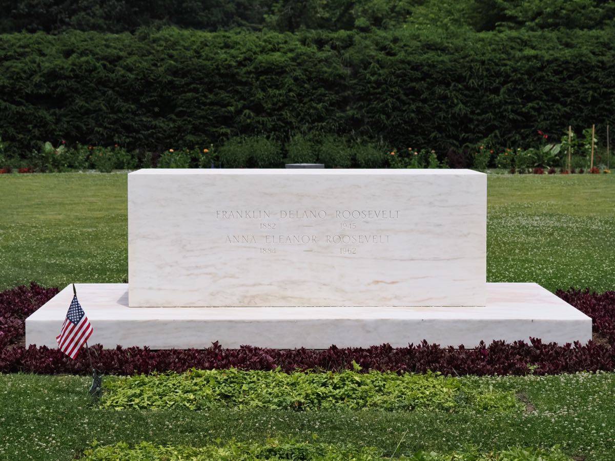 Photo of the grave stone of Franklin and Eleanor Roosevelt at the Roosevelt home in Hyde Park National Historic Site. A simple low, rectangular white marble marker set upon a white marble base with their names and the dates of their births and deaths.