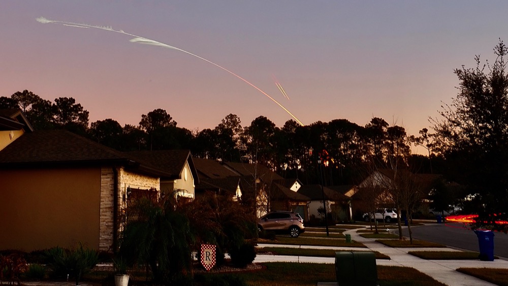 Light trails from the 15 January 2023 Falcon Heavy launched viewed from Ponte Vedra. One light trail arcs from right to left, rising above some trees in a suburban neighborhood setting. Two smaller parallel light trails are above the arc showing the reentry burns of the two side boosters.