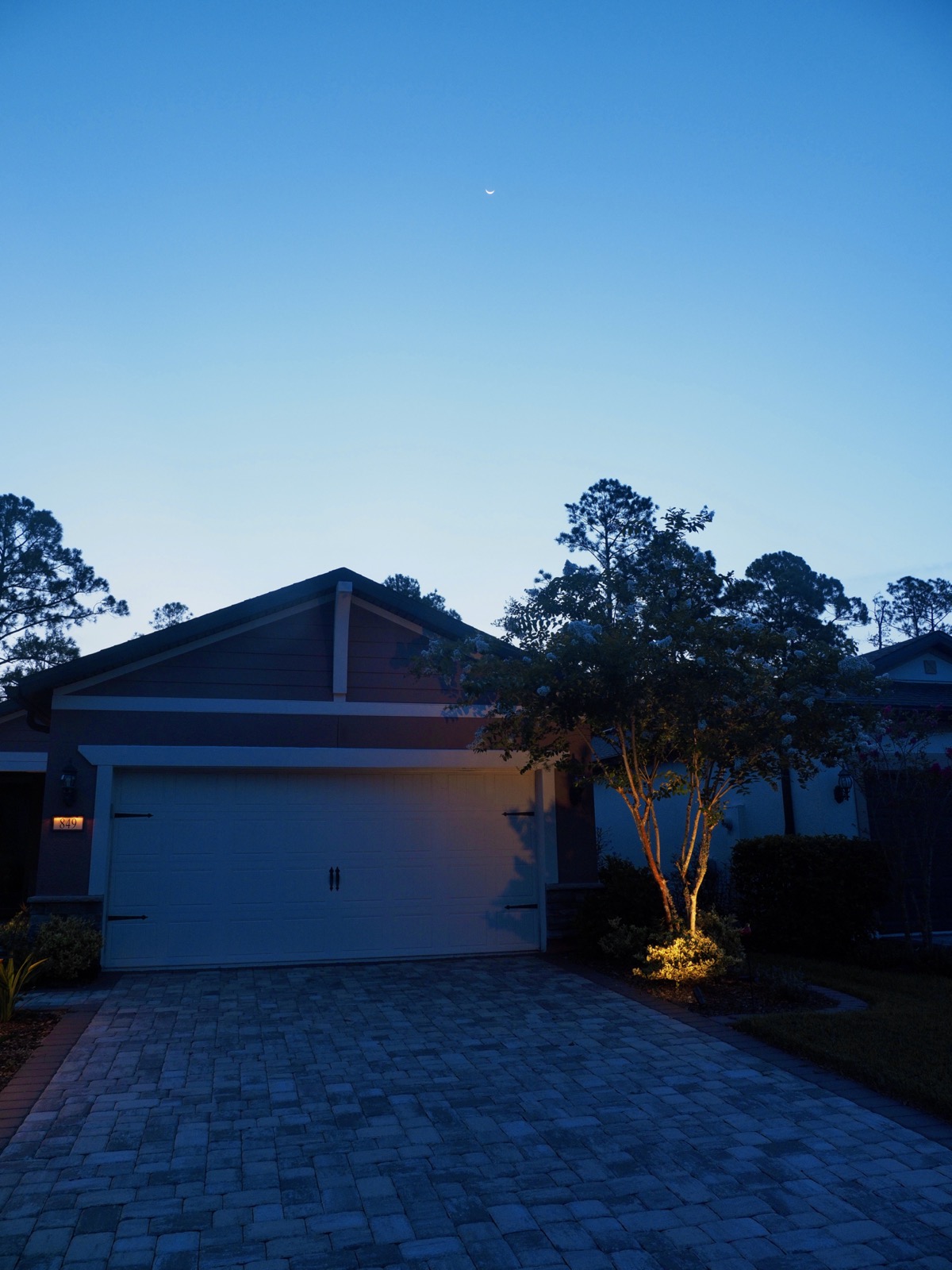 Wide angle portrait orientation shot of a tiny crescent moon barely visible in a blue sky above a suburban house in the minutes before dawn