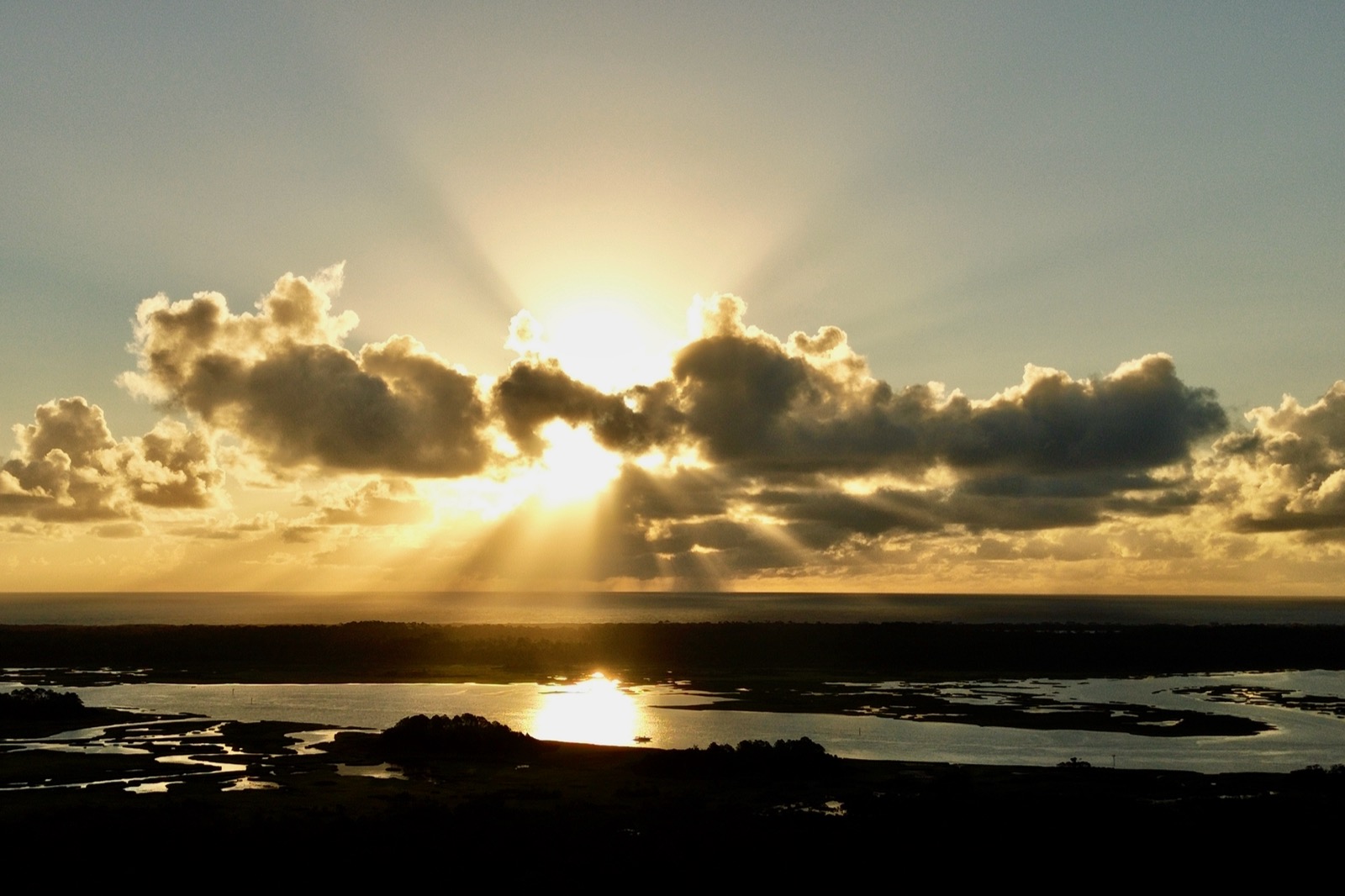 Photo of the early morning sun behind some clouds above the Atlantic Ocean with crepuscular rays eminating and the Tolomato River in the foreground