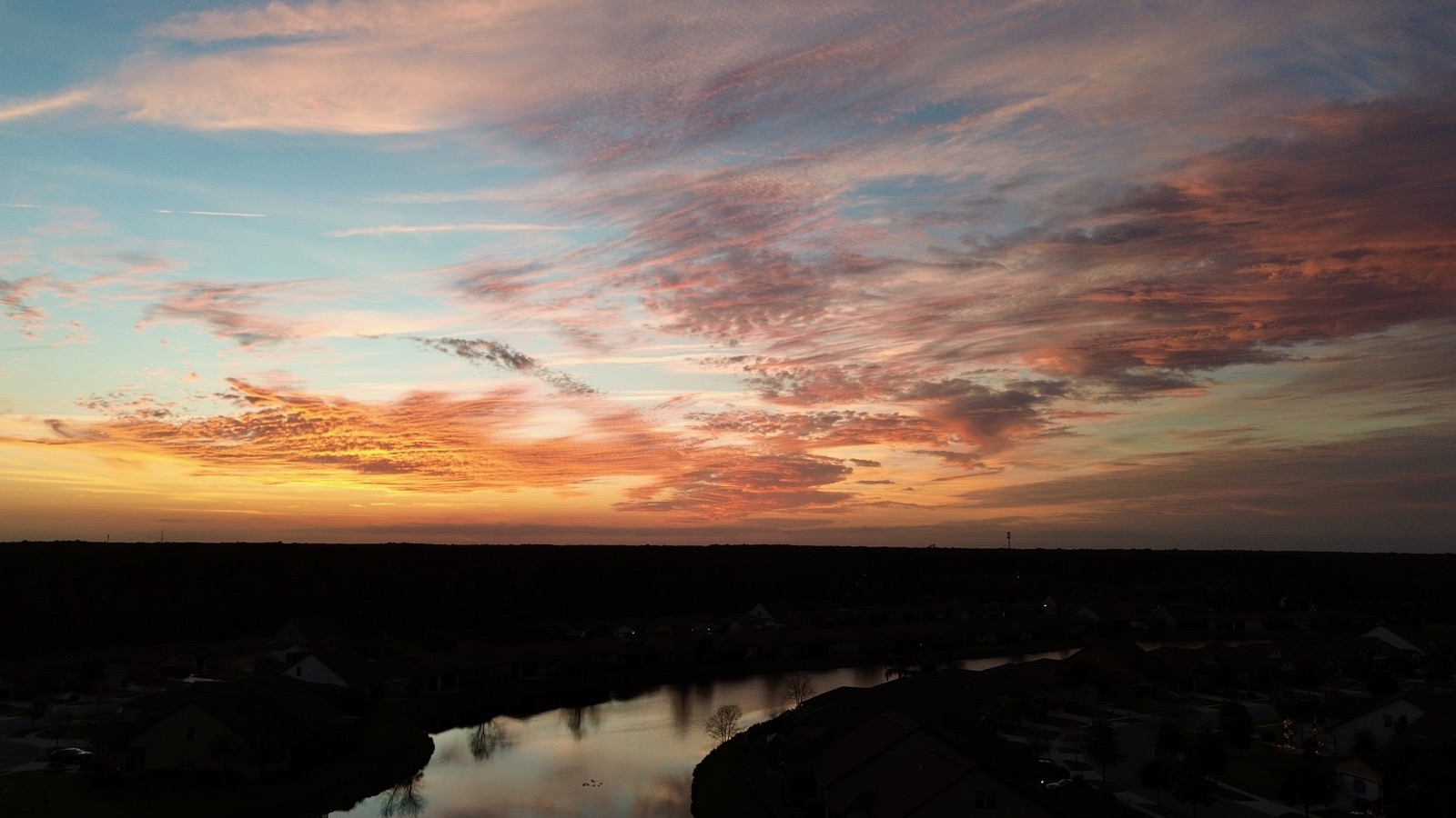 Aerial shot from a drone of clouds illuminated from below by red light from the setting sun, some reflected in a retention pond in the foreground