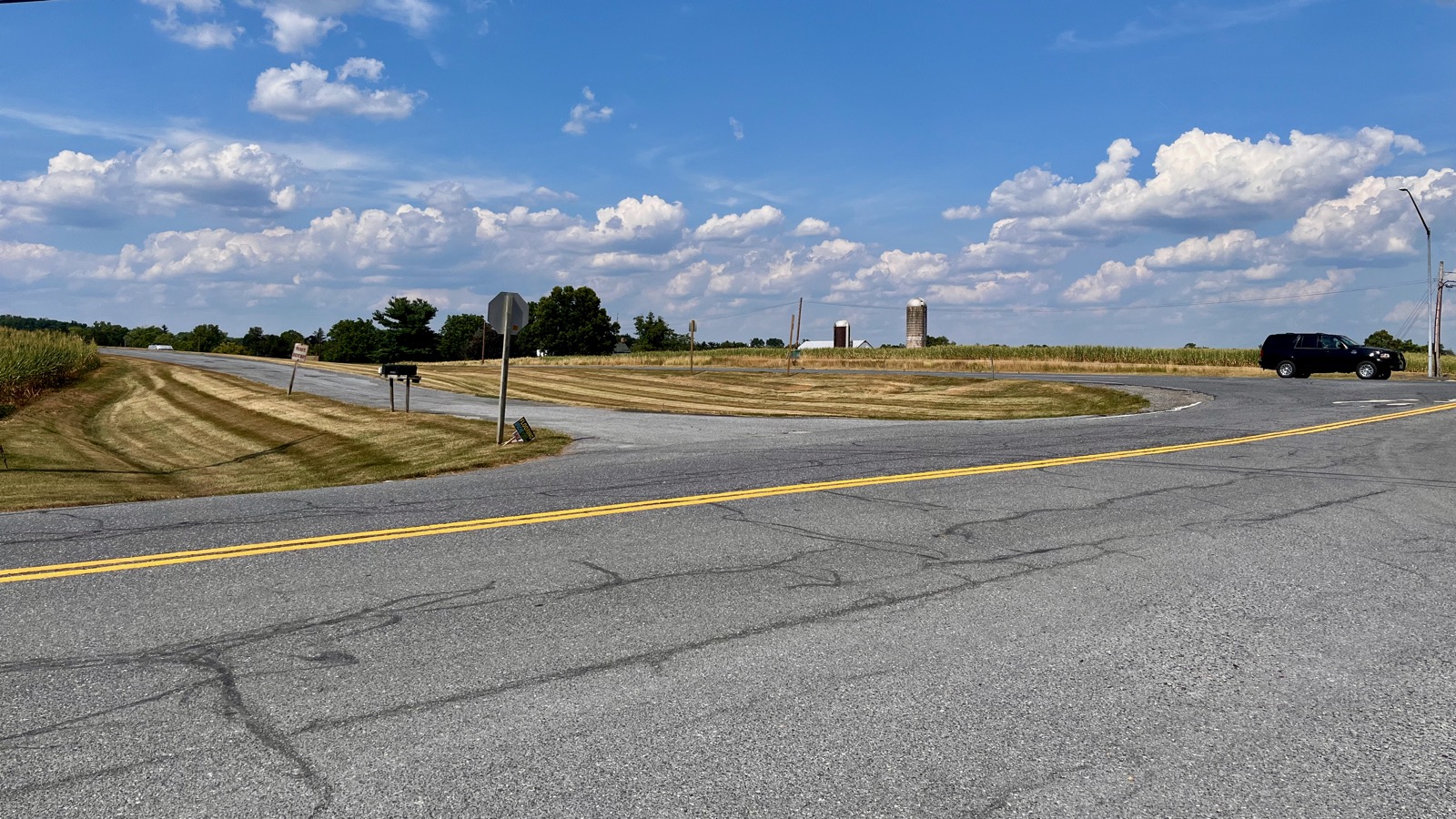 Nondescript bit of asphalt in the foreground, trees, barn and silos in the background with a large SUV at the right edge of the frame