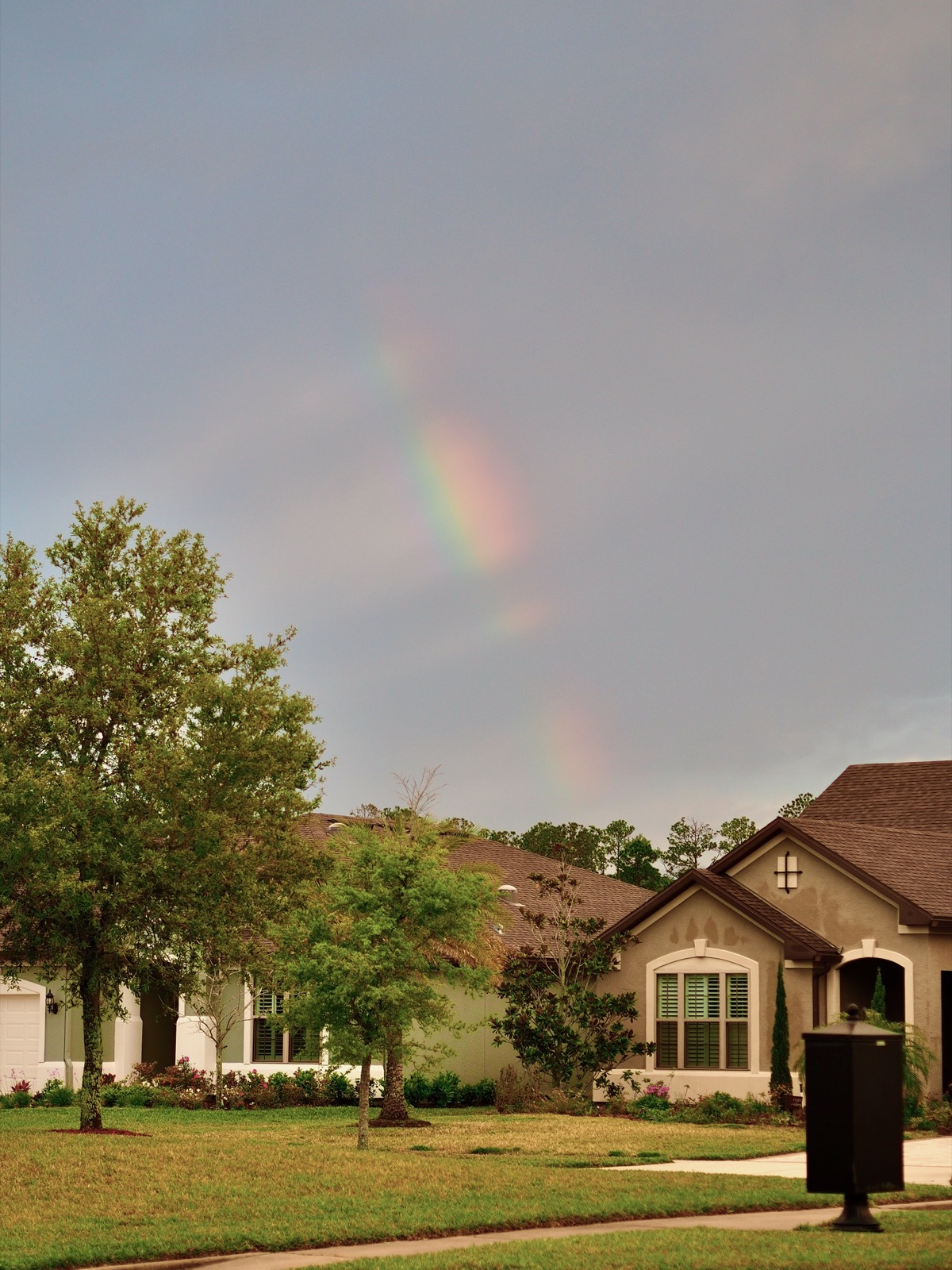 Fragments of a rainbow interrupted by clouds rising from a suburban landscape.