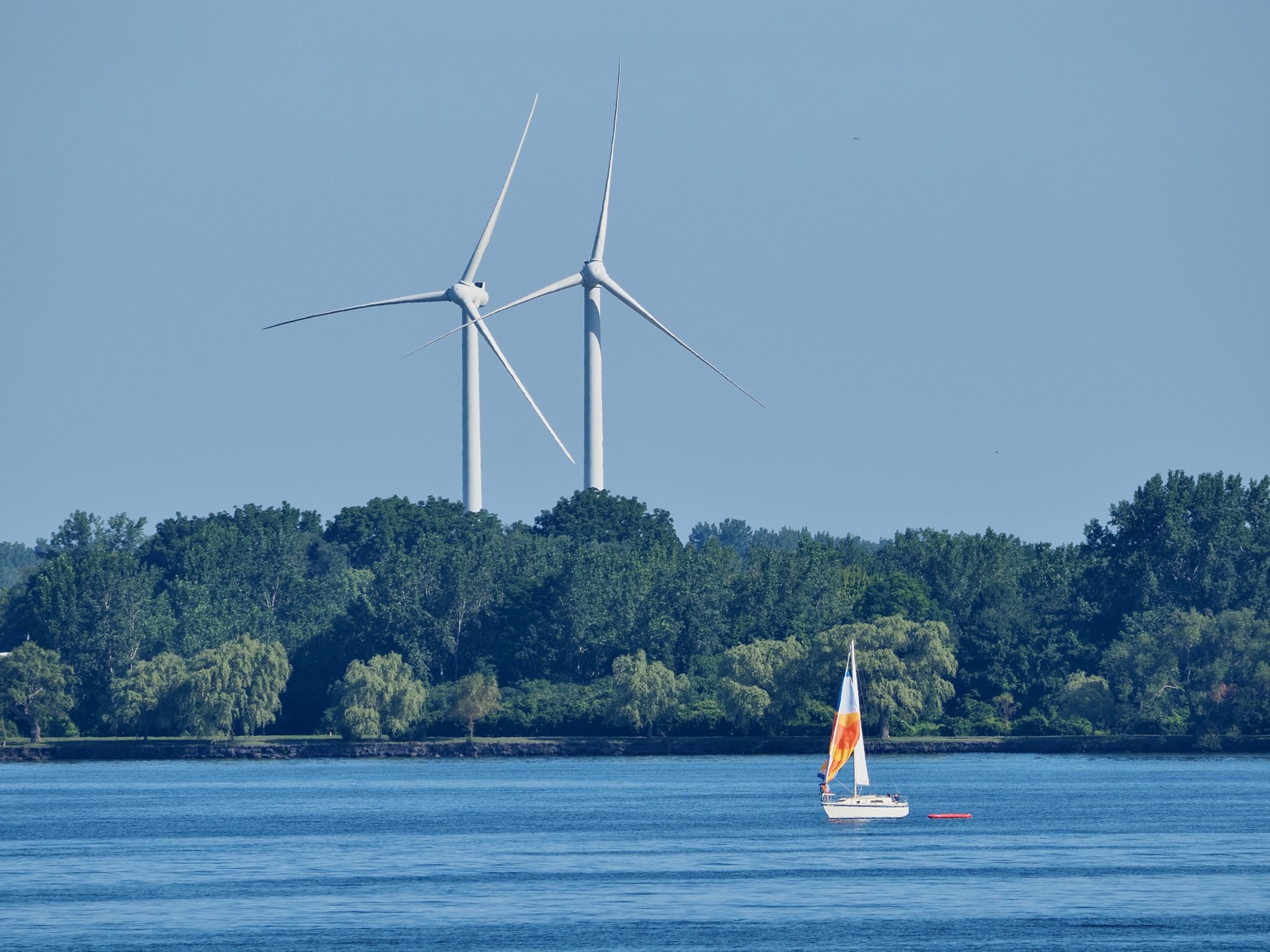 Telephoto image of two large wind turbings in the background towering above some trees along the lakeshore with a sailboat on the lake in the foreground.