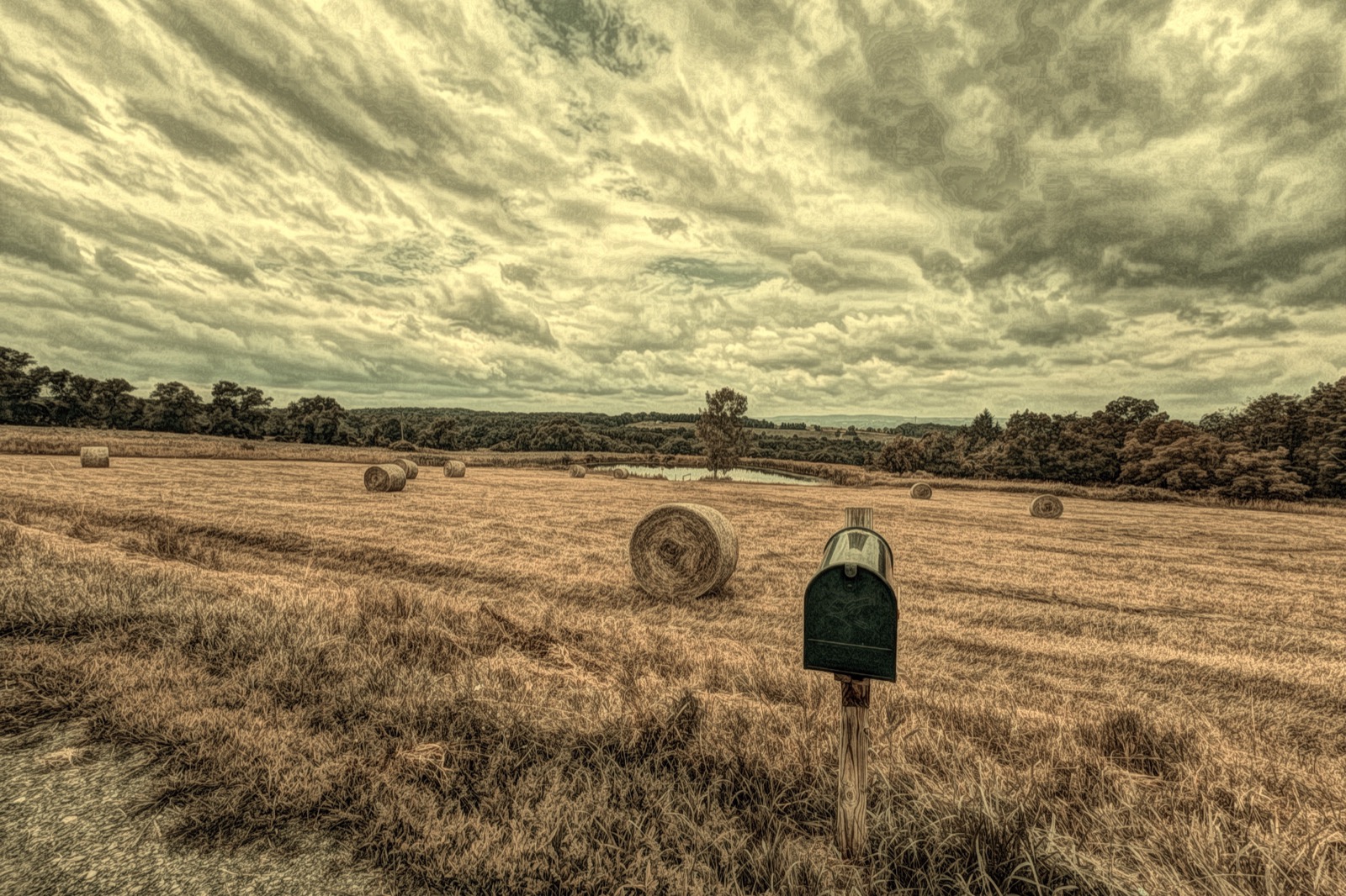 Wide angle image of a hay field with the hay rolled and a mailbox in the foreground edited with a lithograph filter