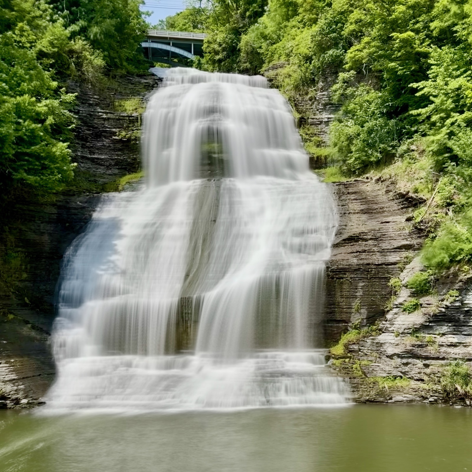 Simulated long-exposure image of a waterfall beneath a bridge, framed on both side by trees and vegetation, terminating in a pond or pool