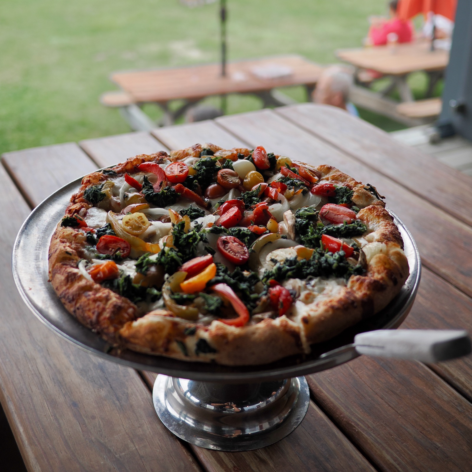 Picture of a pizza on an elevated metal serving platter, set on a woodend picnic table.