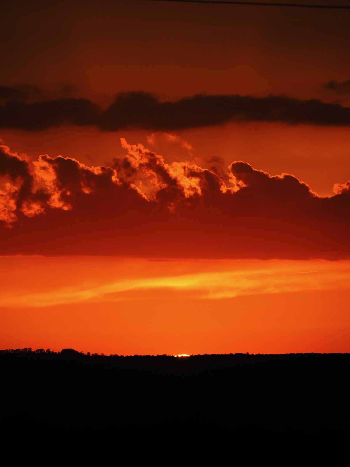 Telephoto image of the upper limb of the setting sun just before it descends below the horizon with and orange dkay and clouds in the background above.