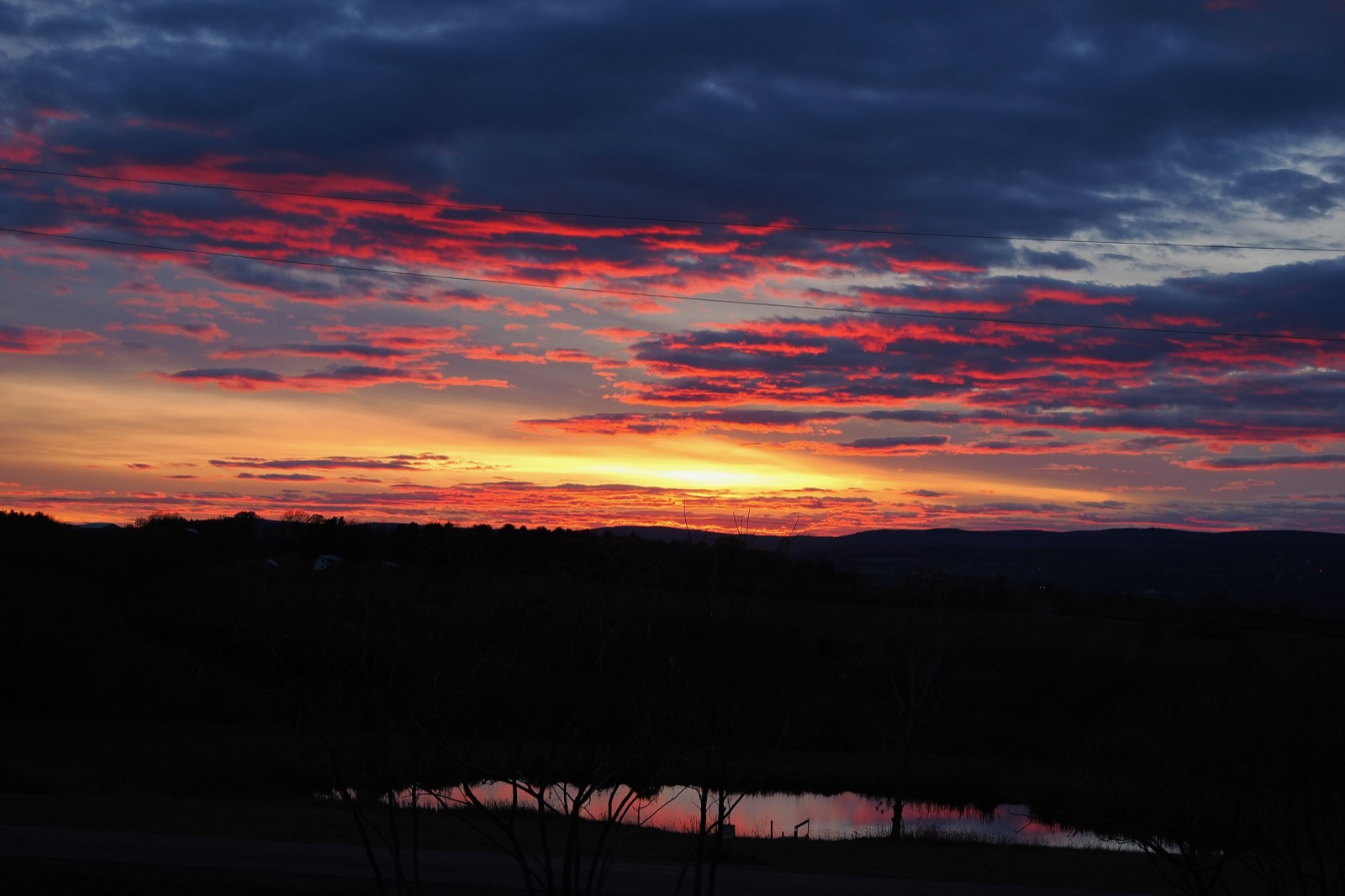 Sunset with broken clouds illuminated in red from the sun below the horizon, a small pond reflects the sky otherwise in shadow.