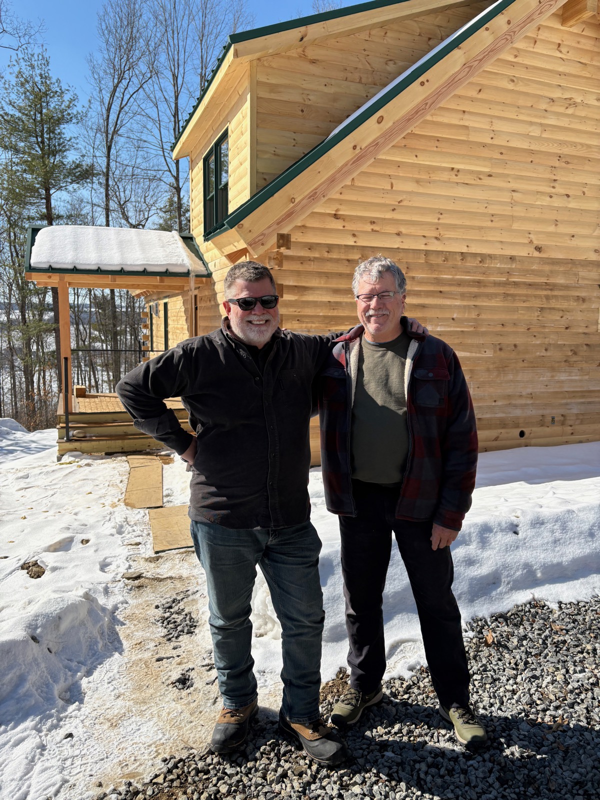 Two old guys standing in the snow in front of a log cabin